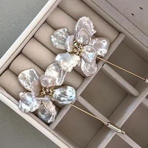 May include: Two floral-shaped brooches with iridescent, irregular-shaped white petals and gold-colored accents. The brooches have long, thin gold-colored pins. The brooches are displayed in a beige jewelry box.