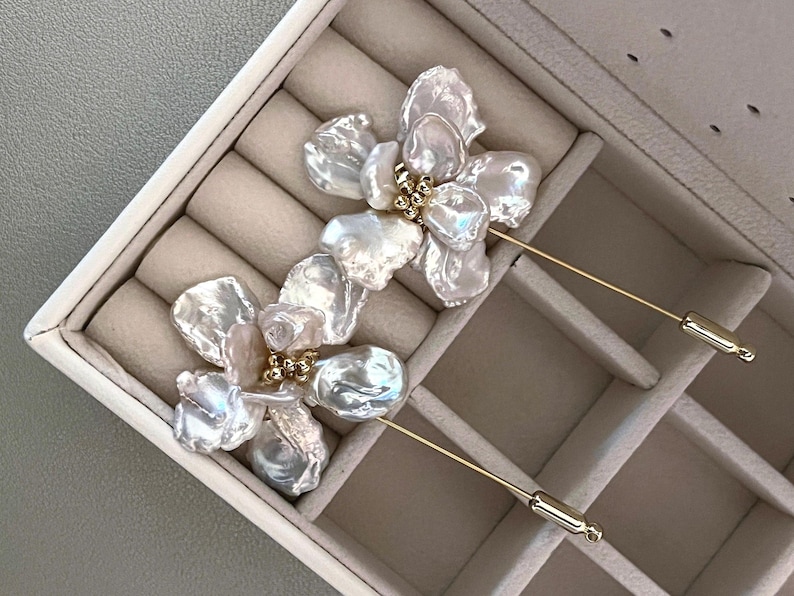 May include: Two floral-shaped brooches with iridescent, irregular-shaped white petals and gold-colored accents. The brooches have long, thin gold-colored pins. The brooches are displayed in a beige jewelry box.