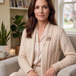 May include: A woman wearing a cream-colored cable-knit cardigan with a matching blouse and pants. A flower brooch adorns the cardigan. The woman is seated in a gray armchair in a well-lit room with a window and a bookshelf.