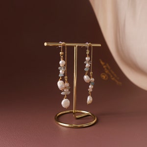 Puede incluir: Un par de pendientes de oro con una cadena delicada y detalles de perlas y piedras preciosas. Los pendientes se muestran en un soporte de oro.