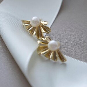 May include: A pair of gold-colored stud earrings with a pearl center. The earrings have a fan-like design. The earrings are displayed on a white ribbon and a gray surface.