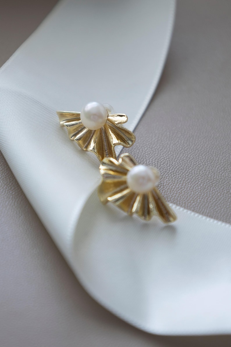 May include: A pair of gold-colored stud earrings with a pearl center. The earrings have a fan-like design. The earrings are displayed on a white ribbon and a gray surface.