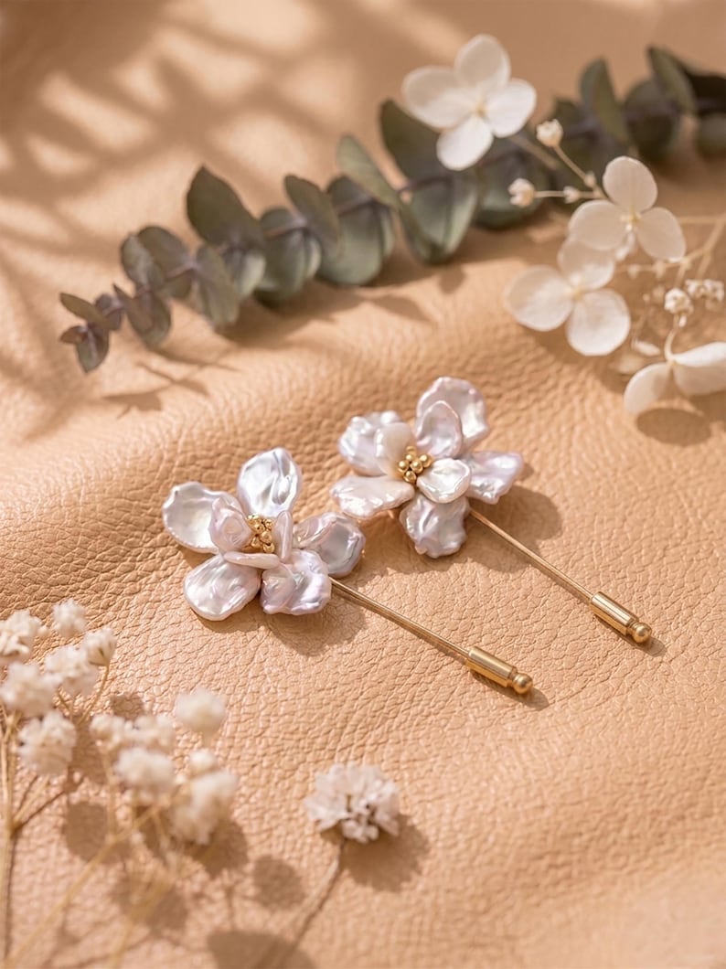 May include: Two flower-shaped brooches with pearl-like petals and gold-colored pin stems. The brooches are set against a tan leather background, with sprigs of white flowers and green leaves scattered around them. The brooches have a delicate, feminine aesthetic.