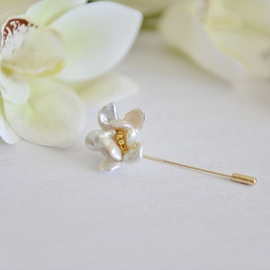May include: A floral-shaped lapel pin with a gold-tone pin and a flower made of white and cream-colored petals. The center of the flower has a cluster of small gold beads. The pin is set against a white background with a blurred flower.