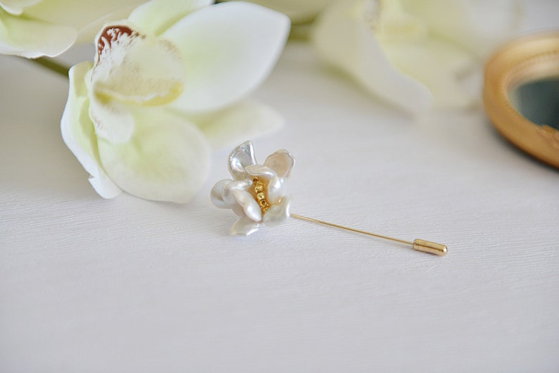May include: A floral-shaped lapel pin with a gold-tone pin and a flower made of white and cream-colored petals. The center of the flower has a cluster of small gold beads. The pin is set against a white background with a blurred flower.