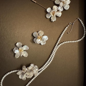 May include: A collection of pearl jewelry featuring floral designs. The set includes a double-strand pearl necklace with a flower pendant, two flower-shaped brooches, and two gold-toned stick pins with flower accents. The jewelry is displayed on a brown surface.