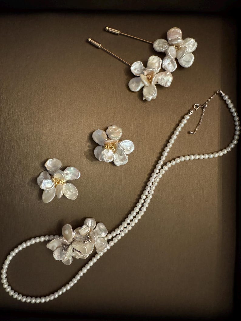 May include: A collection of pearl jewelry featuring floral designs. The set includes a double-strand pearl necklace with a flower pendant, two flower-shaped brooches, and two gold-toned stick pins with flower accents. The jewelry is displayed on a brown surface.