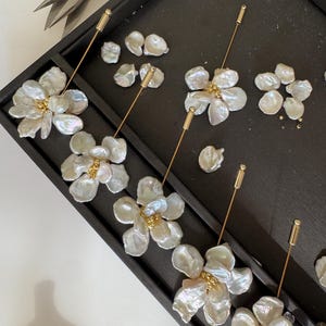 May include: Close-up of handmade floral earrings with iridescent white petals and gold accents. The earrings are arranged on a black tray with additional pearl components and jewelry tools, suggesting a crafting process.
