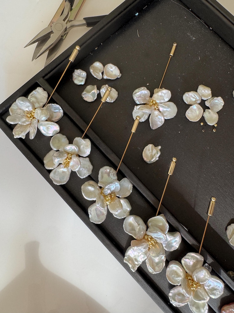 May include: Close-up of handmade floral earrings with iridescent white petals and gold accents. The earrings are arranged on a black tray with additional pearl components and jewelry tools, suggesting a crafting process.