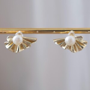 May include: A pair of gold-colored stud earrings. Each earring features a central white pearl set within a fan-shaped, textured gold design. The earrings are displayed on a gold bar against a soft, neutral background.