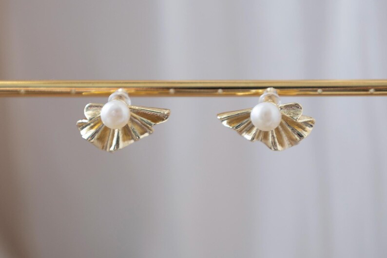 May include: A pair of gold-colored stud earrings. Each earring features a central white pearl set within a fan-shaped, textured gold design. The earrings are displayed on a gold bar against a soft, neutral background.