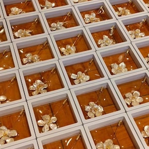 May include: An overhead shot displays numerous floral brooches, each presented in individual white boxes with a wooden-toned interior. The brooches feature a delicate, layered petal design with gold-colored accents, and a long pin.