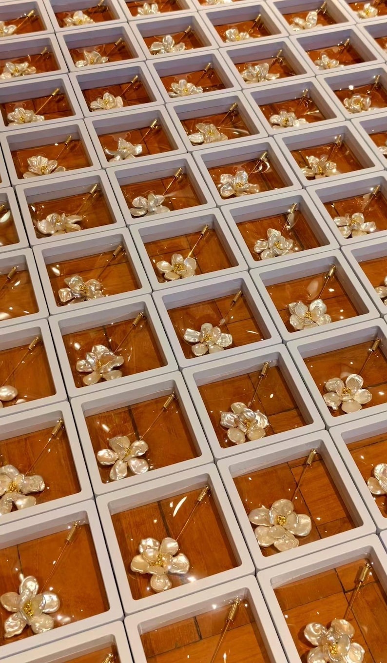 May include: An overhead shot displays numerous floral brooches, each presented in individual white boxes with a wooden-toned interior. The brooches feature a delicate, layered petal design with gold-colored accents, and a long pin.