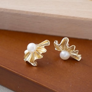 May include: A pair of gold-colored stud earrings. Each earring features a white pearl set in a stylized, ruffled design. The earrings are displayed on a brown surface, with a wooden element in the background.