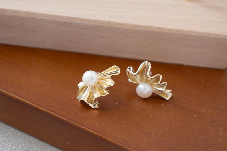 May include: A pair of gold-colored stud earrings. Each earring features a white pearl set in a stylized, ruffled design. The earrings are displayed on a brown surface, with a wooden element in the background.