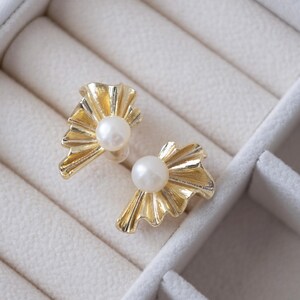 May include: A pair of gold-colored earrings with a pearl center. The earrings have a fan-like design with a textured surface. The earrings are displayed in a white jewelry box.