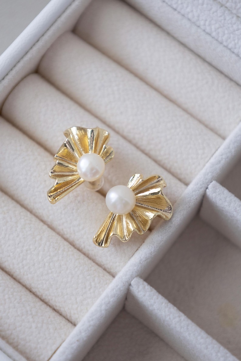 May include: A pair of gold-colored earrings with a pearl center. The earrings have a fan-like design with a textured surface. The earrings are displayed in a white jewelry box.