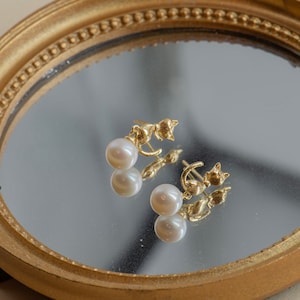 May include: A pair of gold cat-shaped earrings with pearl drops. The earrings are reflected in a gold-framed mirror.