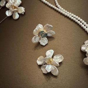 May include: Close-up of elegant jewelry pieces, including flower-shaped earrings and a pearl necklace. The earrings feature delicate, petal-like designs with a central accent. The necklace is composed of small, round pearls. The jewelry is set against a brown background.
