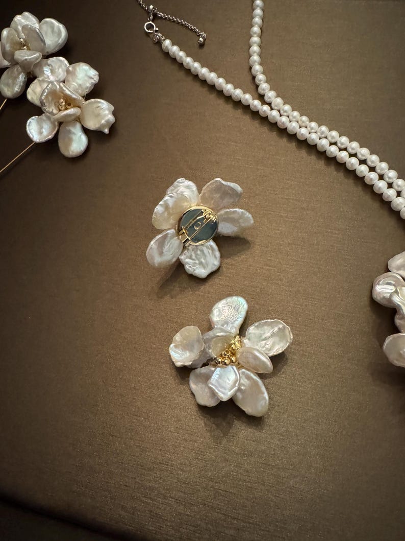 May include: Close-up of elegant jewelry pieces, including flower-shaped earrings and a pearl necklace. The earrings feature delicate, petal-like designs with a central accent. The necklace is composed of small, round pearls. The jewelry is set against a brown background.