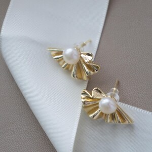 May include: A pair of gold-colored stud earrings with a pearl center and a fan-shaped design. The earrings are displayed on a white ribbon and a neutral-toned surface.