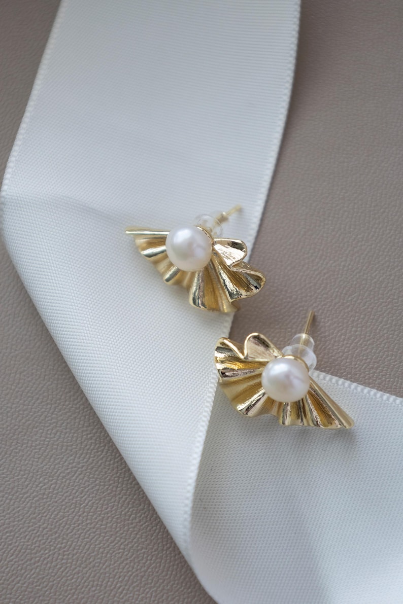 May include: A pair of gold-colored stud earrings with a pearl center and a fan-shaped design. The earrings are displayed on a white ribbon and a neutral-toned surface.