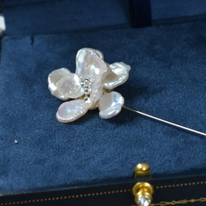 May include: A floral-shaped brooch with a pin, featuring irregular, off-white petals. The center of the flower has small, sparkling embellishments. The brooch is set against a dark blue velvet background.