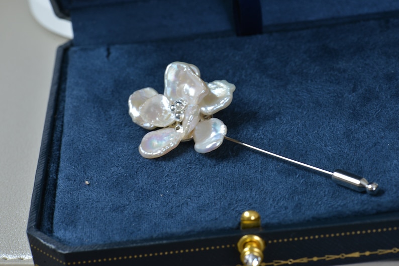 May include: A floral-shaped brooch with a pin, featuring irregular, off-white petals. The center of the flower has small, sparkling embellishments. The brooch is set against a dark blue velvet background.
