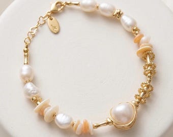 Op de afbeelding: Een goudkleurige armband met witte zoetwaterparels, kleine perzikkleurige schelpschijfjes en gestructureerde goudkleurige metalen tussenstukjes. De armband heeft een karabijnsluiting, een klein ovaal metalen label en een grote, met draad omwikkelde parel.