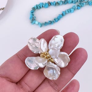 May include: A handcrafted flower-shaped pendant made of large, iridescent white pearls with a gold-colored center. A turquoise bead necklace and small scissors are in the background.