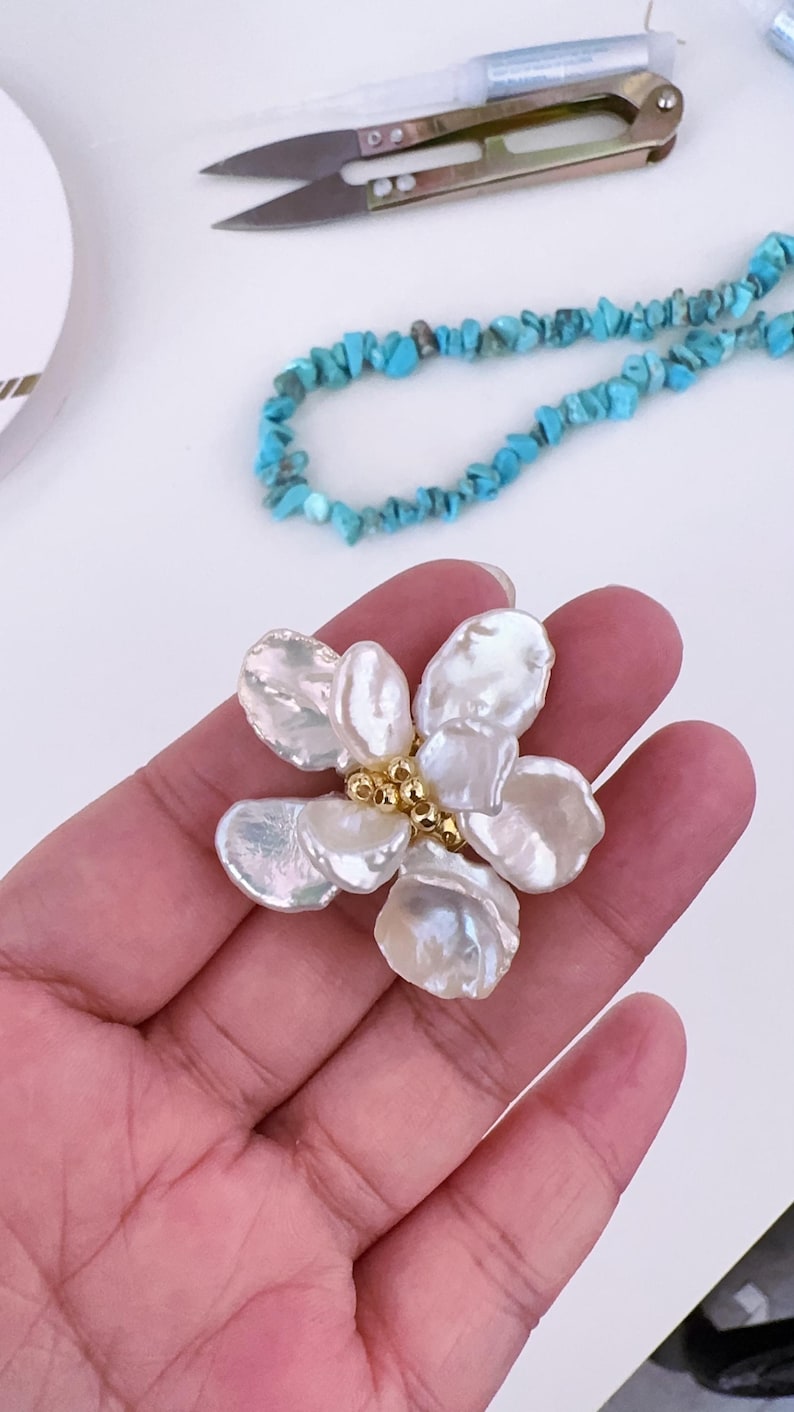 May include: A handcrafted flower-shaped pendant made of large, iridescent white pearls with a gold-colored center. A turquoise bead necklace and small scissors are in the background.