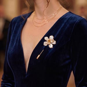 May include: A woman wearing a navy blue velvet dress with a plunging neckline. A gold necklace and a flower-shaped brooch with a gold pin are visible. The brooch has a white and cream color scheme.