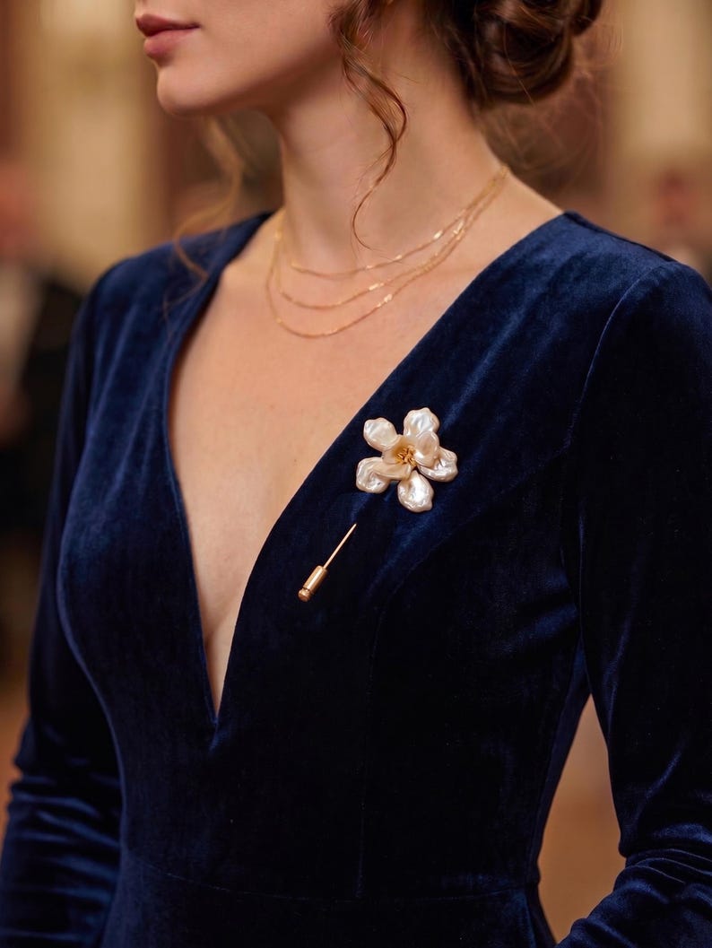May include: A woman wearing a navy blue velvet dress with a plunging neckline. A gold necklace and a flower-shaped brooch with a gold pin are visible. The brooch has a white and cream color scheme.