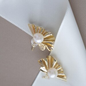 May include: A pair of gold-colored stud earrings. Each earring features a fan-shaped design with a central, round, white pearl. The earrings are set against a white ribbon and a neutral background.