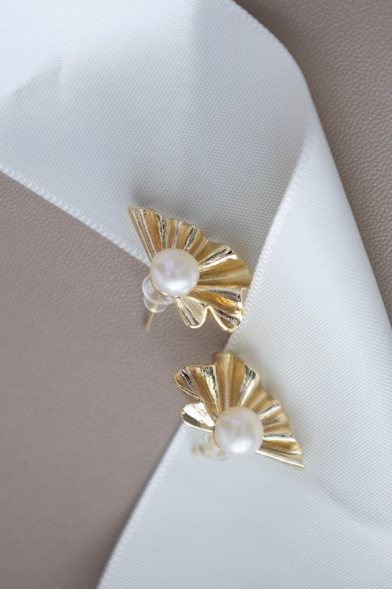 May include: A pair of gold-colored stud earrings. Each earring features a fan-shaped design with a central, round, white pearl. The earrings are set against a white ribbon and a neutral background.