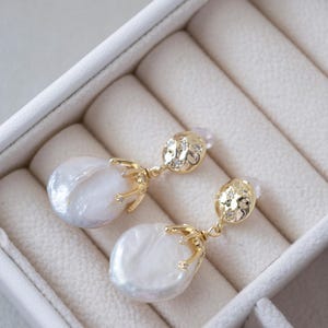 May include: A pair of gold earrings with a white baroque pearl drop and a small gold crown detail. The earrings are in a white velvet jewelry box.
