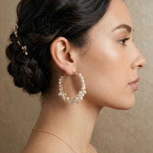 Puede incluir: Pendientes de aro dorados adornados con racimos de perlas blancas. Los pendientes tienen un diseño delicado, perfecto para ocasiones especiales. La imagen muestra un primer plano de los pendientes en una oreja.