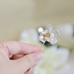May include: A delicate floral brooch featuring a cluster of small gold beads at the center of the flower. The petals are white and have a natural, organic shape. The brooch is held by a hand.