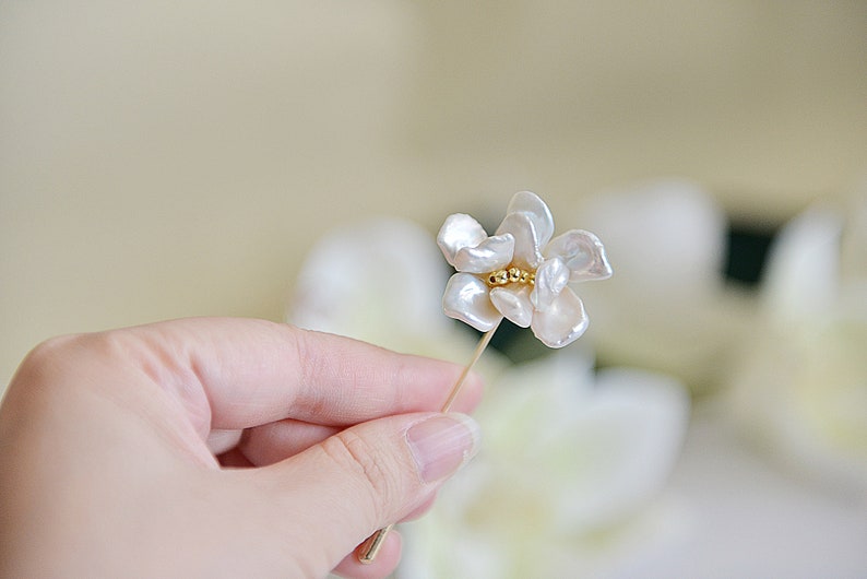 May include: A delicate floral brooch featuring a cluster of small gold beads at the center of the flower. The petals are white and have a natural, organic shape. The brooch is held by a hand.