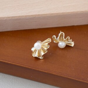 May include: A pair of gold-colored stud earrings. Each earring features a white pearl at the center, surrounded by a stylized, textured gold design. The earrings are displayed on a brown surface.