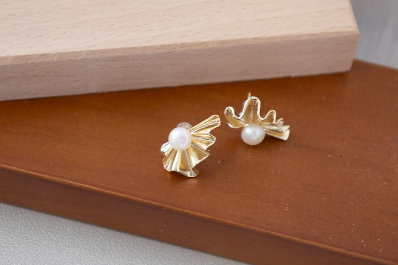 May include: A pair of gold-colored stud earrings. Each earring features a white pearl at the center, surrounded by a stylized, textured gold design. The earrings are displayed on a brown surface.