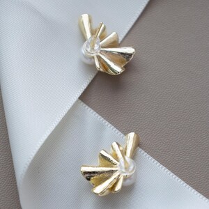 May include: Two gold-colored stud earrings with a fan-like design and a small pearl in the center. The earrings are placed on a white ribbon against a neutral background. The earrings are a jewelry item.