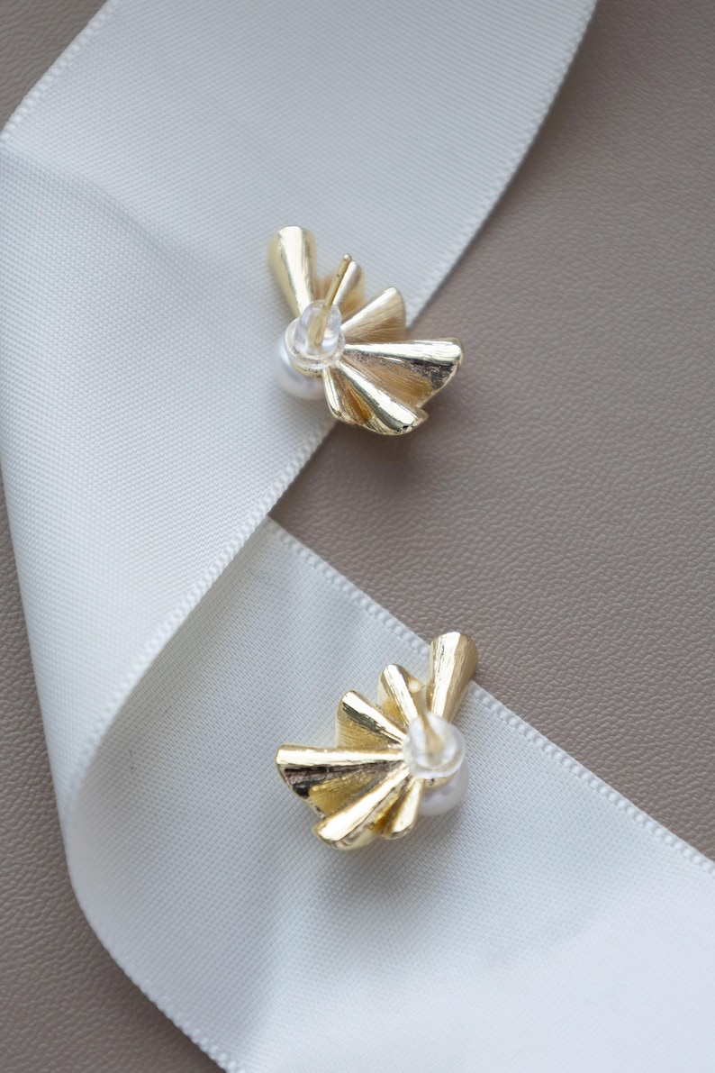 May include: Two gold-colored stud earrings with a fan-like design and a small pearl in the center. The earrings are placed on a white ribbon against a neutral background. The earrings are a jewelry item.