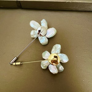 May include: Two flower-shaped brooches. Each brooch features a cluster of irregular, off-white pearls arranged in a floral pattern. One brooch has a silver pin, and the other has a gold pin. The brooches are set against a muted green background.