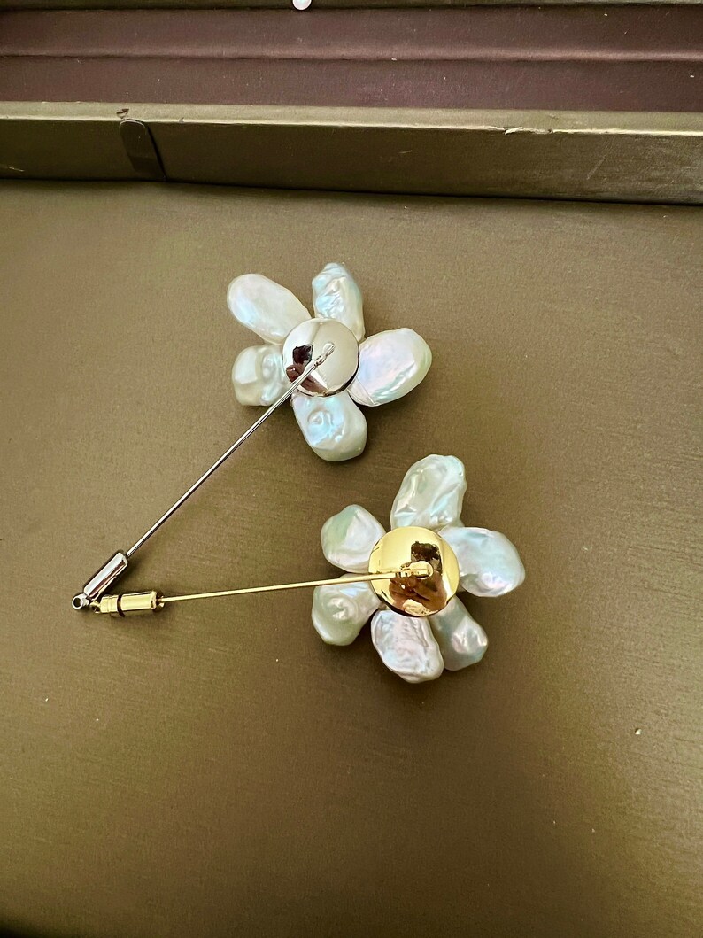 May include: Two flower-shaped brooches. Each brooch features a cluster of irregular, off-white pearls arranged in a floral pattern. One brooch has a silver pin, and the other has a gold pin. The brooches are set against a muted green background.