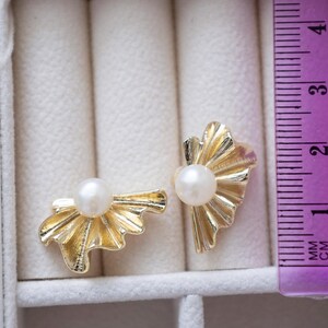 May include: A pair of gold-colored stud earrings with a pearl center. The earrings have a fan-like design. The image includes a ruler showing measurements in centimeters.