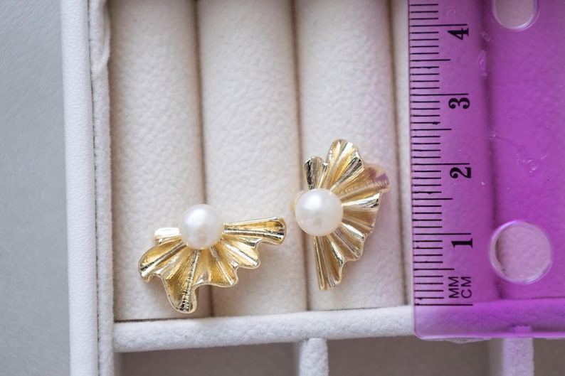 May include: A pair of gold-colored stud earrings with a pearl center. The earrings have a fan-like design. The image includes a ruler showing measurements in centimeters.