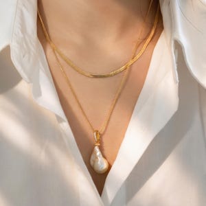 May include: A gold necklace set comprising two layers. The upper layer is a fine chain, and the lower layer features a pendant with a large, irregular pearl. The pendant hangs from a delicate gold chain. The jewellery is worn over a white collared shirt.