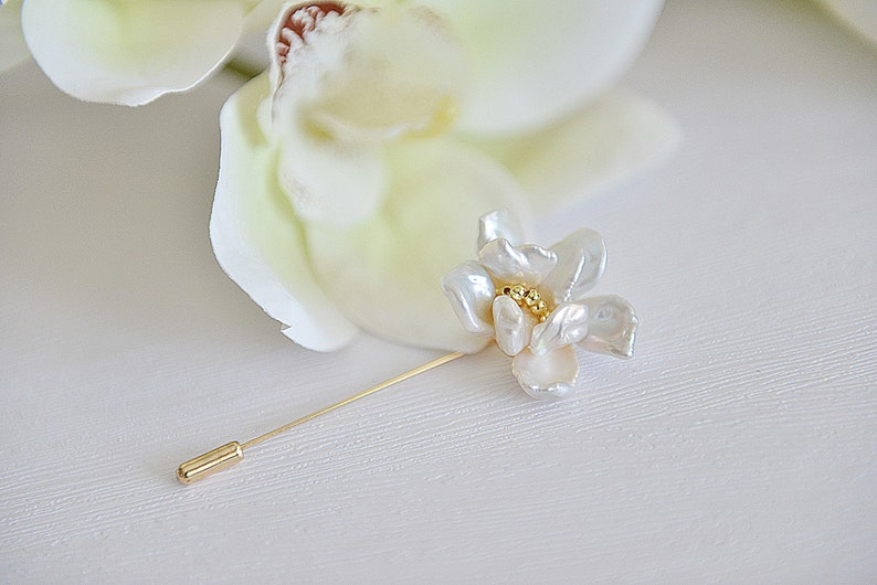 May include: A floral lapel pin featuring a white flower with a gold center and a long, slender gold pin. The flower has a pearl-like texture. The pin is set against a white background, suggesting a wedding or formal occasion.