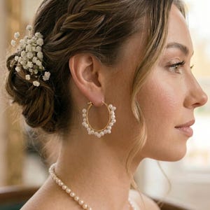 May include: Gold hoop earrings featuring small, white pearls. The earrings are a classic design, adding a touch of elegance. The woman is wearing a pearl necklace and has her hair styled in an updo with a floral hair accessory.